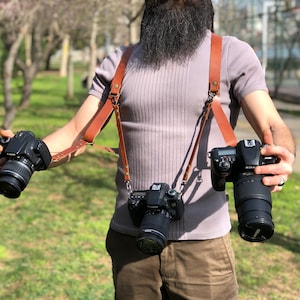 May include: A person wearing a brown leather camera strap with three cameras attached. The cameras are black and have lenses. The person is wearing a grey shirt and brown trousers.