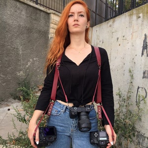May include: A woman with long red hair wearing a black shirt and blue jeans is holding three cameras with red leather camera straps.