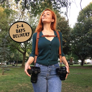 May include: A woman wearing a green cardigan and blue jeans is using a brown leather camera strap to carry two cameras. The strap is worn across her chest and has a buckle at the front. The text "2-4 DAYS DELIVERY" is in a circle in the top left corner of the image.