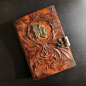 May include: A brown leather journal with a decorative metal clasp and a Native American chief head embossed on the front cover.