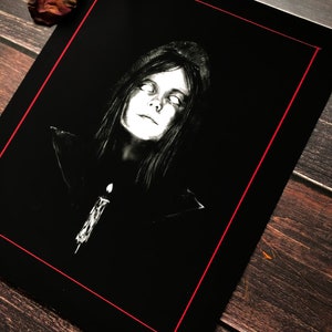 Puede incluir: Ilustraci&oacute;n en blanco y negro de una persona con ojos brillantes y una vela. La obra de arte est&aacute; enmarcada con un borde rojo y sobre un fondo oscuro. La persona lleva una prenda con capucha.