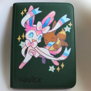 Puede incluir: Una carpeta de color verde oscuro con cremallera que presenta una ilustración gráfica del personaje Sylveon rosa y blanco junto a un personaje Eevee marrón. La portada muestra el texto vaultx en letras negras, acentuado por estrellas amarillas.