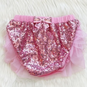 May include: Pink sequined baby bloomers with a pink bow and a ruffled tulle trim. The bloomers have an elastic waistband and are designed for infants. The sequins shimmer, adding a touch of sparkle to the garment.