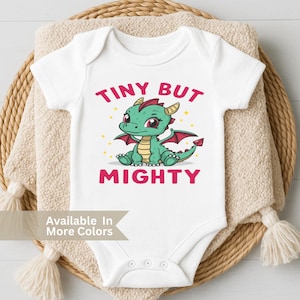 May include: White baby onesie with a cartoon dragon illustration. The dragon is green with red accents and the text "TINY BUT MIGHTY" in pink. The onesie is displayed on a beige blanket and a woven basket.