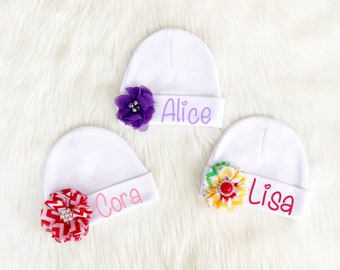 May include: Three white baby hats with different colored flowers and names embroidered on them. The names are Alice, Cora, and Lisa.