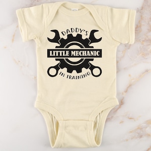 May include: A cream-colored baby onesie with a black graphic of a gear and two wrenches. The text on the onesie reads "Daddy's Little Mechanic In Training".