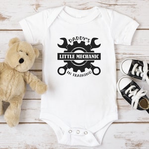 May include: White baby onesie with black text and graphic. The text reads "Daddy's Little Mechanic in Training" with a gear and wrench design.