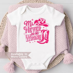 May include: White baby bodysuit with a pink graphic that reads "Ms. Never Do No Wrong" with a cowboy hat and boots. The bodysuit is displayed on a pink blanket.
