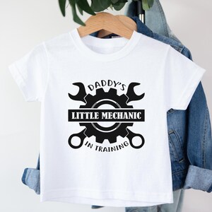 May include: White t-shirt with a black graphic of a gear and wrenches. The text reads "Daddy's Little Mechanic in Training".