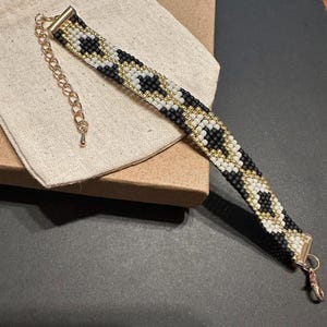 May include: A beaded bracelet with a black, white, and gold pattern. The bracelet has a gold-colored clasp and chain for adjusting the size. The bracelet is resting on a beige fabric and a cardboard box.