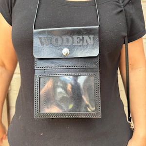 May include: Black leather crossbody bag with the word "WODEN" embossed on the flap. The bag has a clear ID window and a snap closure. The strap is thin and black.
