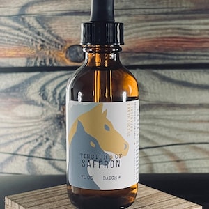 Könnte beinhalten: Eine braune Glasflasche mit Tropfer und einem weißen Etikett. Das Etikett zeigt eine stilisierte Illustration eines Pferdekopfes und den Text "Tincture of Saffron" mit den Wörtern "FL OZ" und "BATCH #" darunter. Die Flasche steht auf einer Holzoberfläche.