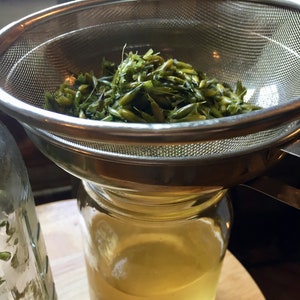 May include: A stainless steel strainer with green plant material inside, being used to strain a clear liquid into a glass jar.