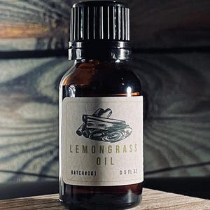 Pode incluir: Uma pequena garrafa de vidro castanho de óleo de capim-limão com uma tampa preta. O rótulo apresenta o texto "LEMONGRASS OIL" e um gráfico. A garrafa está sobre um bloco de madeira, com um fundo de madeira escura.