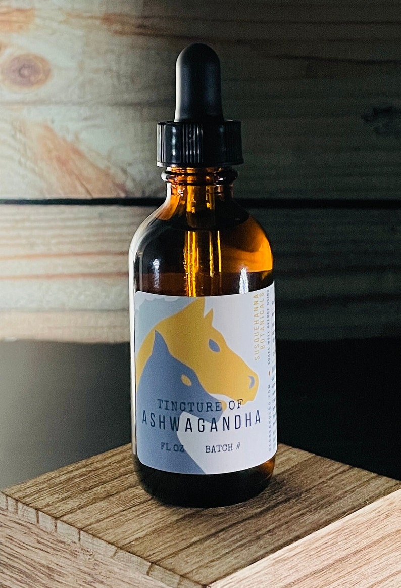 Op de afbeelding: Een bruine glazen fles met een druppelaar en een etiket met de tekst "Tincture of Ashwagandha" met een gele en blauwe illustratie van een paardenkop. De fles staat op een houten oppervlak.