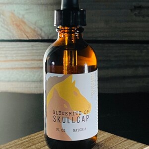 Può includere: Una bottiglia di vetro marrone con un contagocce, etichettata "Glycerite of Skullcap" e "FL OZ BATCH #". La bottiglia è appoggiata su una superficie di legno.