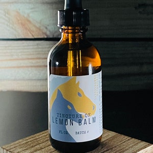 May include: Amber glass bottle with a black dropper, labeled "Tincture of Lemon Balm." The label features a blue and yellow horse design. The bottle is on a wooden surface, with a wooden plank background. The product is from Susquehanna Botanicals.