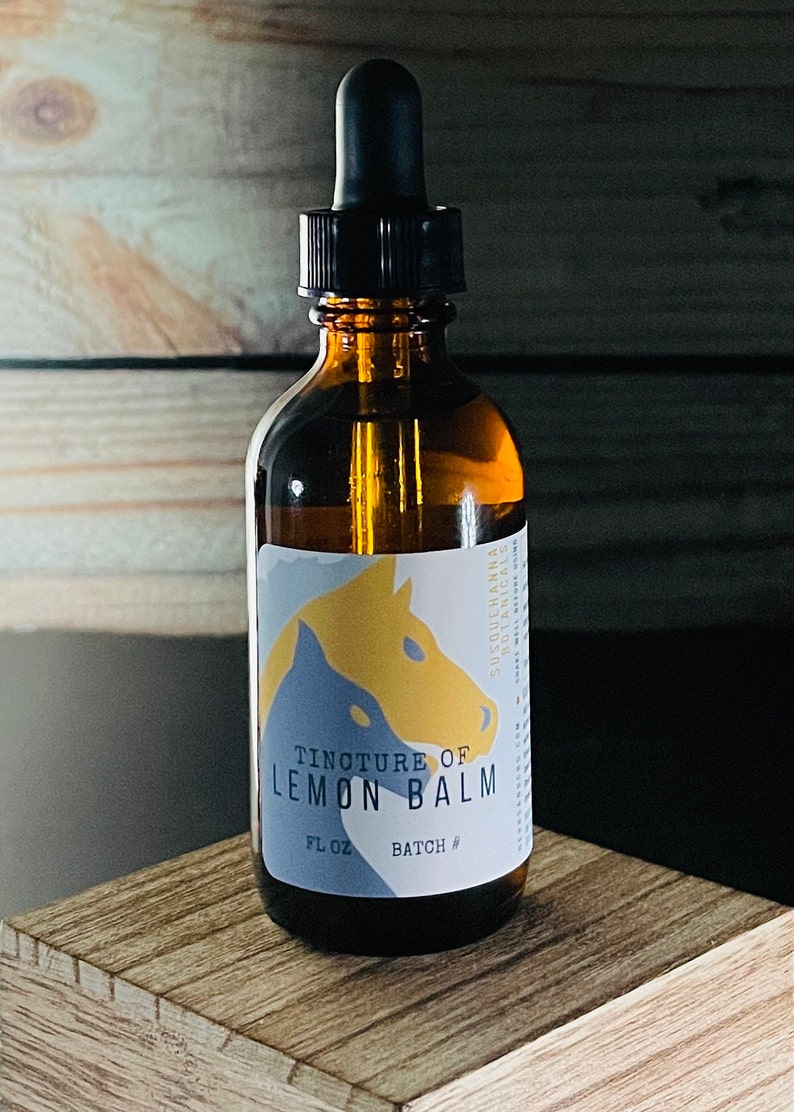 May include: Amber glass bottle with a black dropper, labeled "Tincture of Lemon Balm." The label features a blue and yellow horse design. The bottle is on a wooden surface, with a wooden plank background. The product is from Susquehanna Botanicals.
