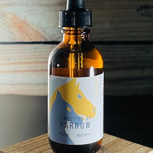 Puede incluir: Una botella de vidrio marrón con un gotero, etiquetada "Tintura de Milenrama" con una ilustración de un caballo amarillo y azul. La botella está sobre una superficie de madera.