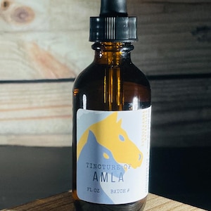 Puede incluir: Una botella de vidrio marrón con un gotero y una etiqueta que dice "Tincture of Amla" con una ilustración amarilla y azul de una cabeza de caballo. La botella está sobre una superficie de madera.