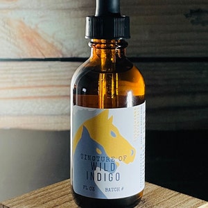Op de afbeelding: Een bruine glazen fles met een druppelaar en een etiket met de tekst "Tincture of Wild Indigo" en "FL OZ BATCH #". De fles staat op een houten oppervlak.
