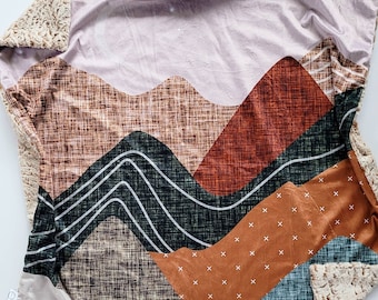Puede incluir: Una manta suave y mullida con un diseño geométrico de montaña. La manta presenta una mezcla de colores marrón, beige, naranja, verde y blanco. La tela tiene un aspecto texturizado y tejido.
