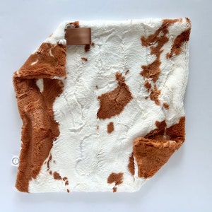 May include: A soft, plush blanket with a brown and white cow print design. The blanket has a brown satin ribbon attached to one corner.