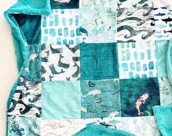 Puede incluir: Una colcha de retazos con cuadrados en tonos azules y blancos. Los cuadrados muestran ballenas, narvales y otras criaturas marinas. La colcha está bordeada con una tela suave de color turquesa.
