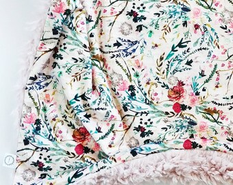 May include: A floral patterned blanket with a cream background and a variety of flowers in pink, red, and teal. The blanket has a soft, fuzzy, light pink border. The blanket is folded, showing the floral pattern and the border.