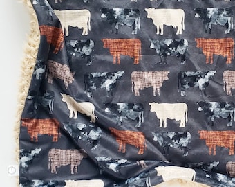 Puede incluir: Una manta suave y mullida con un fondo gris oscuro y un patrón repetitivo de siluetas de vacas marrones, blancas y rojas. La manta tiene un borde peludo de color beige.