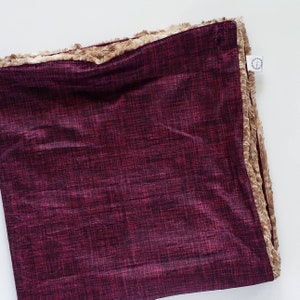 May include: A square, burgundy-colored blanket with a textured pattern. The blanket is trimmed with a soft, tan-colored faux fur. A small white tag is attached to the edge of the blanket.