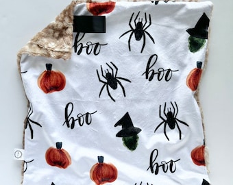 Puede incluir: Una manta de felpa blanca y marrón con temática de Halloween. La manta presenta un patrón repetitivo de arañas negras, calabazas naranjas y la palabra "boo" escrita en negro.