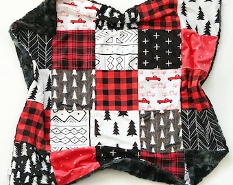 Puede incluir: Una manta de retazos con varios patrones en rojo, negro y blanco. Los diseños incluyen camiones rojos, cuadros escoceses, árboles y formas geométricas. La manta tiene un borde de felpa gris oscuro y suave.