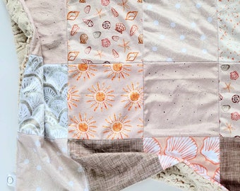 Puede incluir: Una colcha de retazos con una variedad de patrones, incluyendo conchas marinas, soles y diseños geométricos. La colcha está hecha de tela suave de color beige y tiene un respaldo blanco esponjoso.