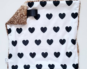 Puede incluir: Una manta blanca para bebé con un patrón de corazones negros. La manta tiene un ribete de piel sintética marrón y una etiqueta negra.
