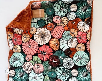 Puede incluir: Una manta cuadrada para bebé con un estampado de calabazas en tonos naranja, rosa, verde y blanco sobre un fondo negro. La manta tiene un borde de felpa marrón y una etiqueta de satén.