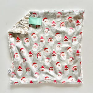 May include: A gray and white Christmas-themed baby blanket with a pattern of Santa Claus faces. The blanket has a soft, plush texture on one side and a smooth, printed fabric on the other. A mint green ribbon is attached to the blanket.