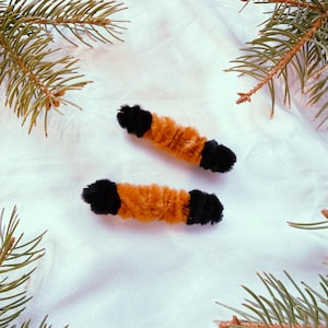 Puede incluir: Dos pinzas para el pelo hechas a mano con limpiapipas naranja y negro. Las pinzas son cilíndricas con extremos negros y un centro naranja. El fondo es blanco con ramas de pino verdes en las esquinas.