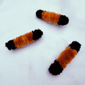 May include: Three orange and black fuzzy hair clips are arranged on a white surface. The clips are cylindrical, with black ends and an orange center. The texture of the clips appears soft and plush.