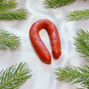 May include: A red sausage shaped Christmas ornament hanging from a white string. The ornament is surrounded by green pine branches.