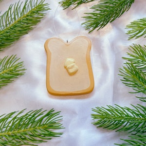 May include: A handmade ceramic ornament shaped like a slice of toast with a pat of butter on top. The ornament is painted in shades of yellow and brown and has a string for hanging.