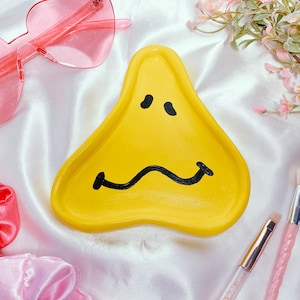 Peut inclure: Plateau en céramique jaune en forme de visage triste avec des traits peints en noir. Le plateau est sur une surface en satin blanc avec des accessoires roses.