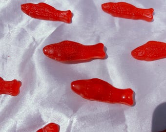 May include: Bright red, fish-shaped gummy candies arranged on a white, textured surface. The candies have a glossy appearance and are scattered across the fabric. A close-up shot highlights the details of the treats.