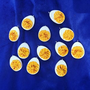 May include: A collection of egg-shaped pendants with a white base and a yellow and orange swirl design, resembling deviled eggs. The pendants are arranged on a blue surface.