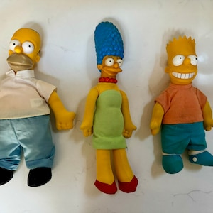 May include: Three yellow plush toys of the Simpsons characters: Homer, Marge, and Bart. Homer is wearing a white shirt and blue jeans. Marge is wearing a green dress. Bart is wearing a pink shirt and blue shorts.