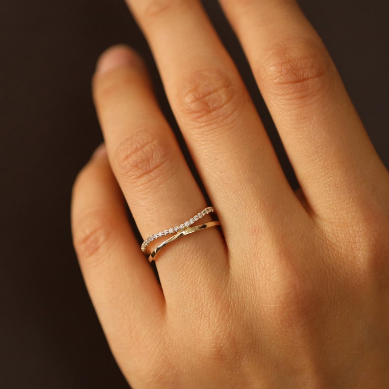 May include: A gold-colored criss-cross ring with a band of small, clear gemstones on one side. The ring is on a person's finger, with a dark background.