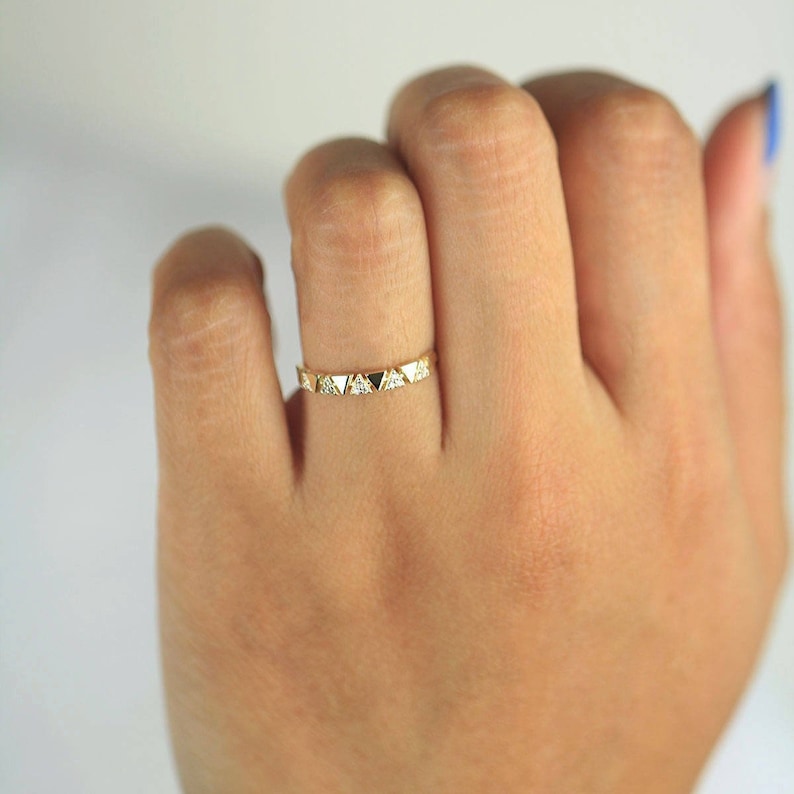 Pu&ograve; includere: Un anello d'oro con un motivo ripetuto di piccoli triangoli, alcuni con pietre trasparenti e altri con pietre nere. L'anello &egrave; indossato su un dito, mettendo in risalto il suo design. Un delicato gioiello.