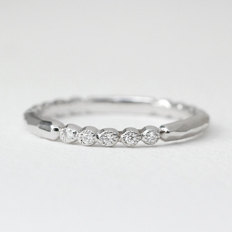 May include: A silver ring with six small, round diamonds set in a row. The band has a textured, faceted design. The ring is displayed against a white background, highlighting its sparkle and elegance.