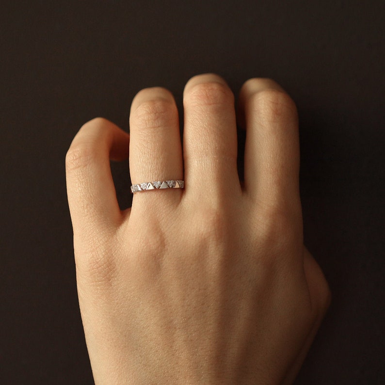 Pu&ograve; includere: Un delicato anello d'argento con un motivo ripetuto di piccoli triangoli e accenti scintillanti. L'anello &egrave; indossato su un dito, mettendo in risalto il suo design intricato e la sua sottile eleganza. Il design dell'anello &egrave; una rivisitazione moderna di uno stile classico.