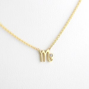 Puede incluir: Un collar de oro con un peque&ntilde;o acento de diamante y un colgante en forma de la letra "M".